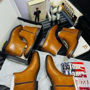 Four pairs of tan leather boots are displayed with designer books, a figurine, and a photo in the background, all arranged on a black slatted surface.