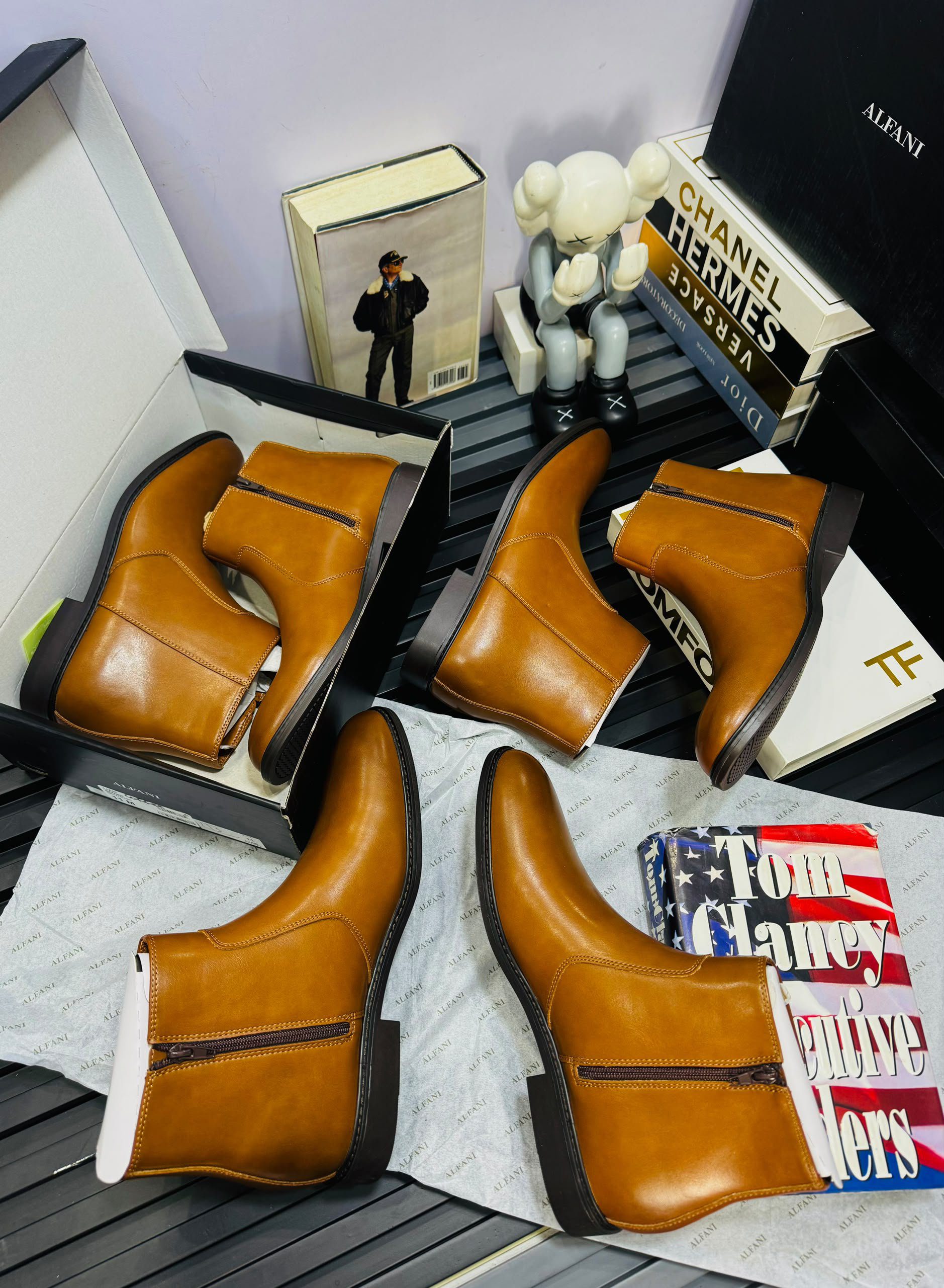 Four pairs of tan leather boots are displayed with designer books, a figurine, and a photo in the background, all arranged on a black slatted surface.